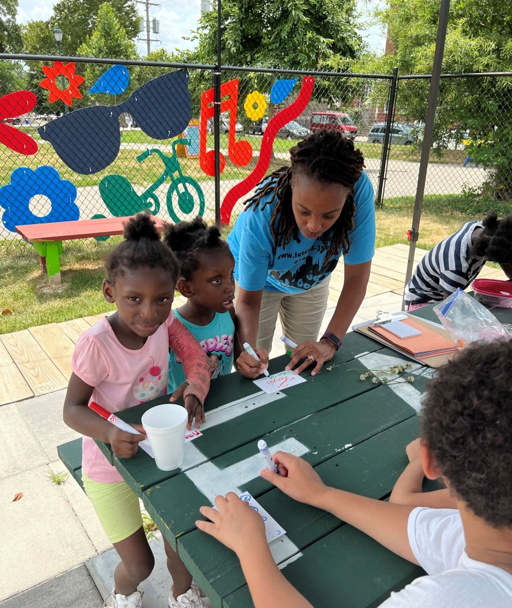 Children work on an activity at a picnic table with an adult guiding them.